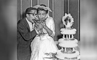 Sammy Davis Jr and Loray White at their wedding drinking from a large glass next to their cake 